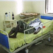 Travel Medicine - Patient in private hospital, notice the sink is not working behind him and family provided sheets and drinks.